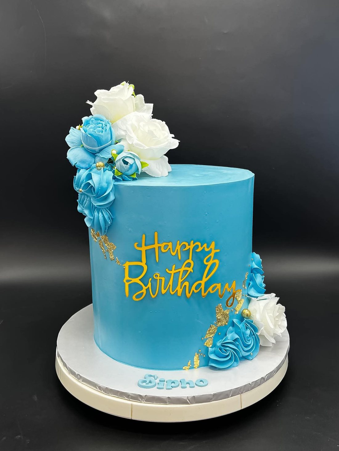 A tall blue buttercream birthday cake with white and blue roses and a gold Happy Birthday topper